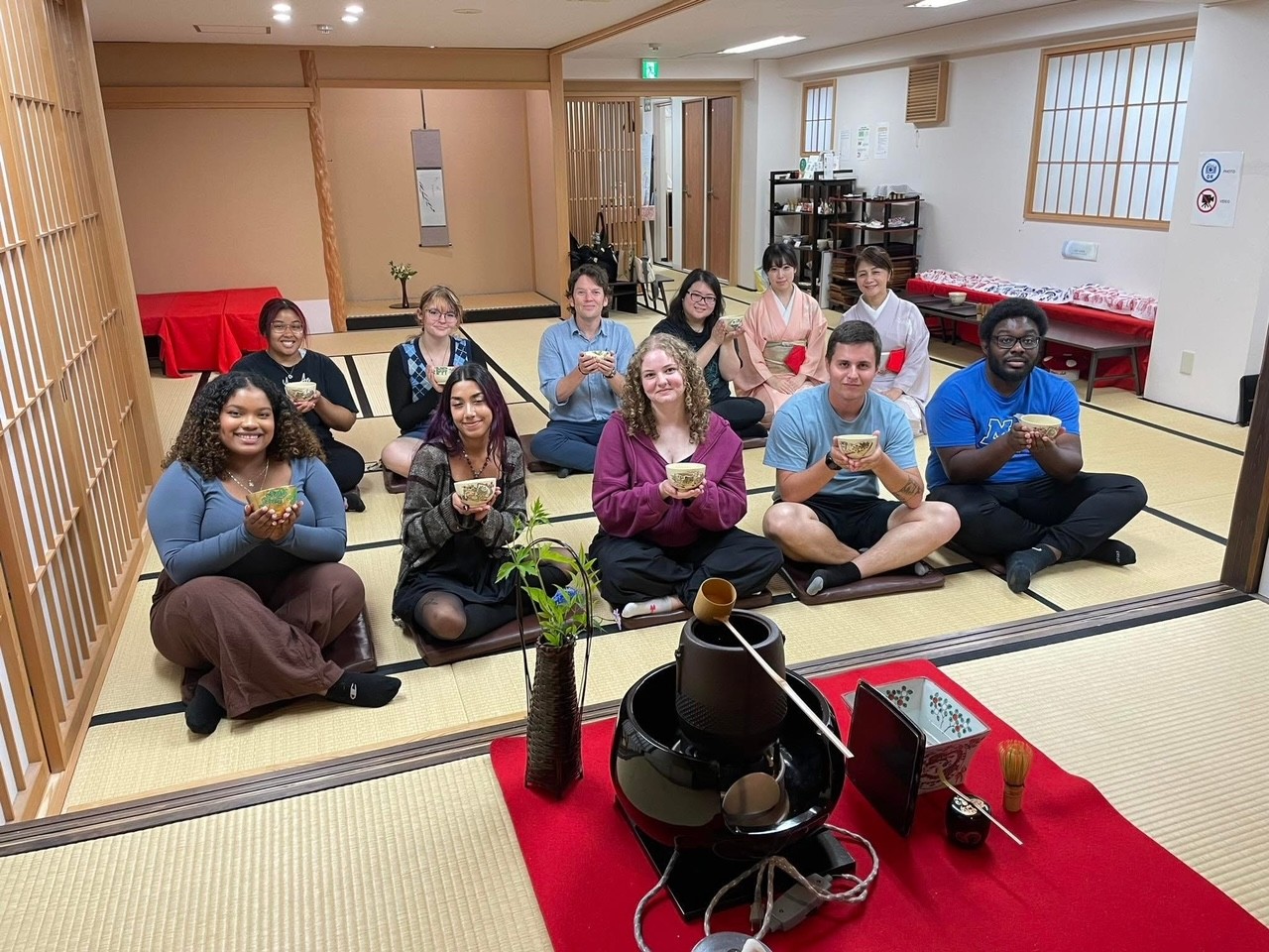 A group of students holding up a cup and sitting on the floor inside a house in Japan.